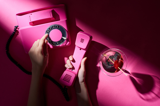 Cropped view of woman using retro telephone with martini beside on pink background - Photo, image