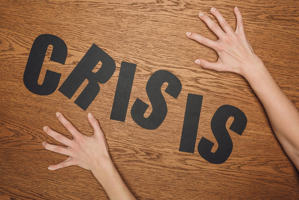 Ausgeschnittene Ansicht weiblicher Hände in der Nähe von schwarzem Papier geschnitten Wort Krise auf Holzboden - Foto, Bild