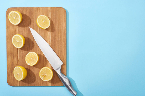 top view of ripe yellow cut lemons on wooden cutting board with knife on blue background with copy space - Foto, Bild