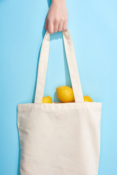 青い背景のコットンバッグに熟した黄色のレモンを持つ女性のクロップドビュー - 写真・画像