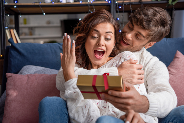 handsome boyfriend giving present to shocked girlfriend in sweater at christmastime - Foto, Bild