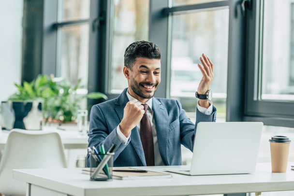 excited businessman showing winner gesture while sitting at workplace near laptop - Photo, Image
