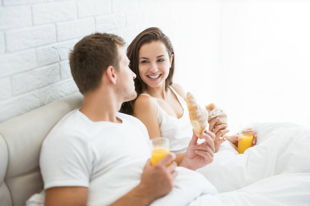 happy young couple handsome man and beautiful woman having breakfast in bed eating croissant and drinking orange juice looking happy morning family idyll - Photo, Image