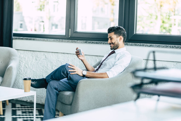 enfoque selectivo de un hombre de negocios sonriente sentado en un sillón y usando un teléfono inteligente
 - Foto, imagen