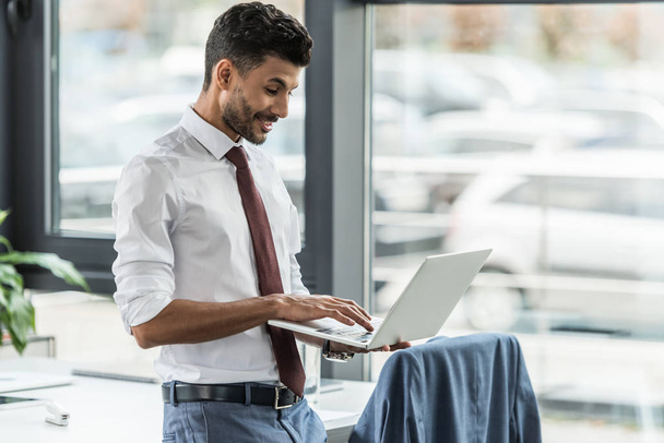 усміхнений бізнесмен використовує ноутбук, стоячи на робочому місці
 - Фото, зображення