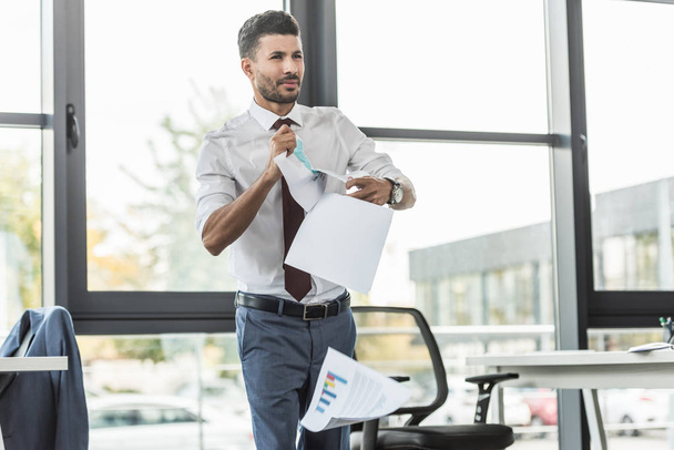 displeased businessman tearing documents and looking away while standing near window in office - Photo, image
