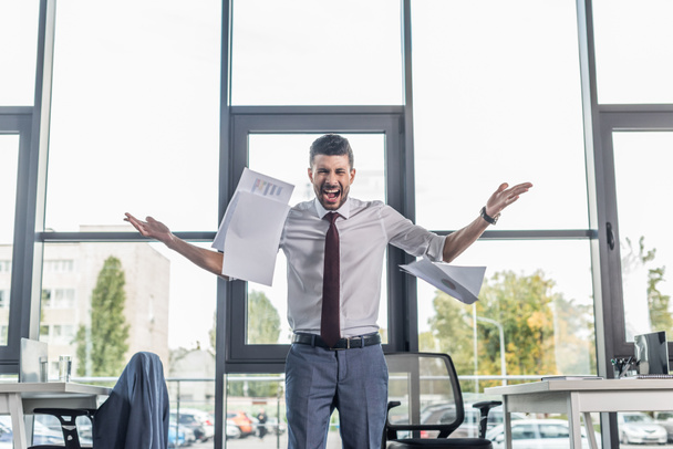 boze zakenman gooien van papieren en schreeuwen terwijl staan in modern kantoor - Foto, afbeelding