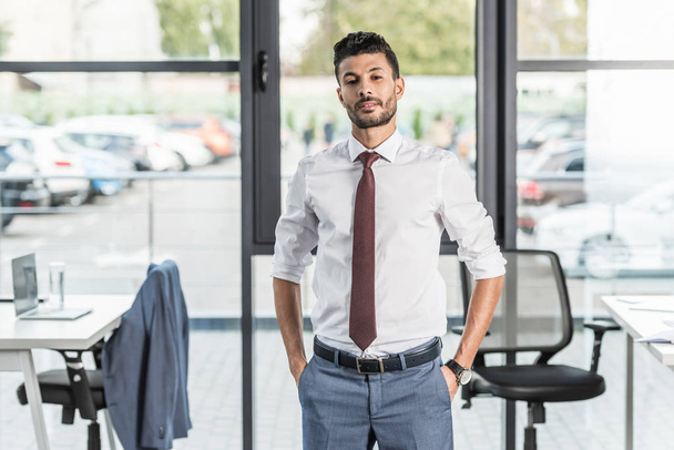 confident businessman looking at camera while holding hands in pockets - Photo, Image
