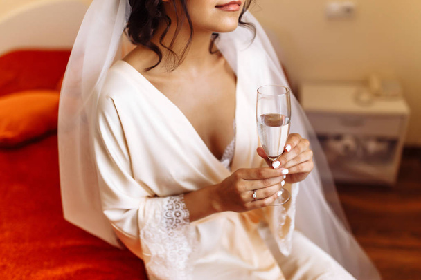 Beautiful bride in a white robe holds in her hand a glass of champagne. Elegant woman with professional make up and hair style. Wedding morning of the bride. Wedding day. Wedding dress. Fashion bride. - Foto, afbeelding