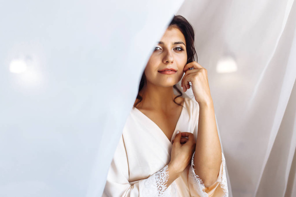 Elegant bride in white robe posing under curtain. Amazing bride in the morning preparation. Beautiful woman with professional make up and hair style. Morning of the bride. Wedding day. Fashion bride. - Фото, изображение