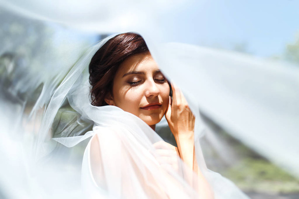 A beautiful portrait of the bride under a veil. Elegant woman with professional make up and hair style. A photo of delicate bride's hands hidden under a veil. Wedding day. Marriage. Fashion bride. - Foto, Imagem