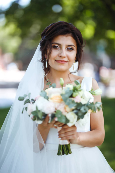Beautiful bride with a wedding bouquet for a walk in the park. Young bride in white dress. Beautiful woman with professional make up and hair style. A beautiful bride portrait. Wedding day. Marriage. - Photo, image