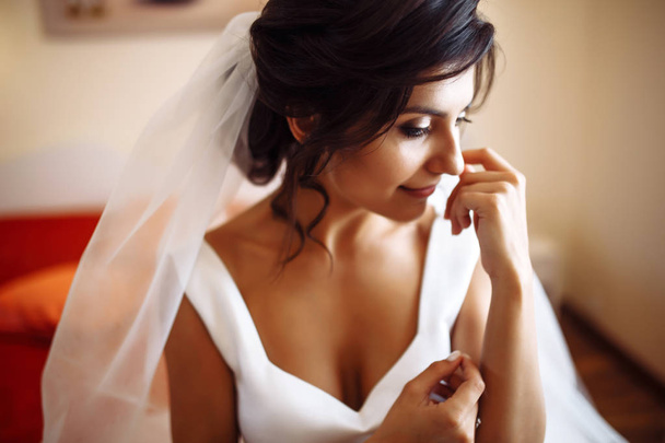 Morning of the bride. Beautiful smiling brunette bride in a white dress with a wedding bouquet in her hands. Elegant woman with professional make up and hair style. Wedding morning of the bride.  - Fotó, kép