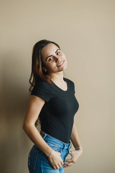 Happy young woman friendly looking to camera at beige background. She clothed in black t-shirt and blue jeans - 写真・画像