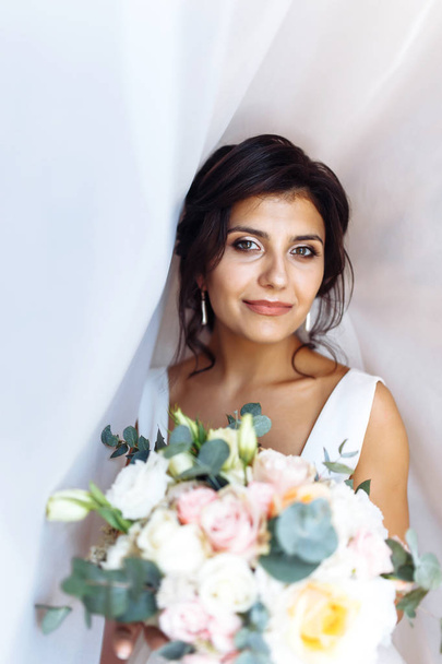 Young bride in white dress with a bouquet of flowers posing under curtain. Beautiful woman with professional make up and hair style. Morning of the bride. Wedding day. Fashion bride. - Φωτογραφία, εικόνα