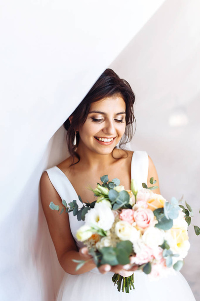 Young bride in white dress with a bouquet of flowers posing under curtain. Beautiful woman with professional make up and hair style. Morning of the bride. Wedding day. Fashion bride. - Фото, зображення