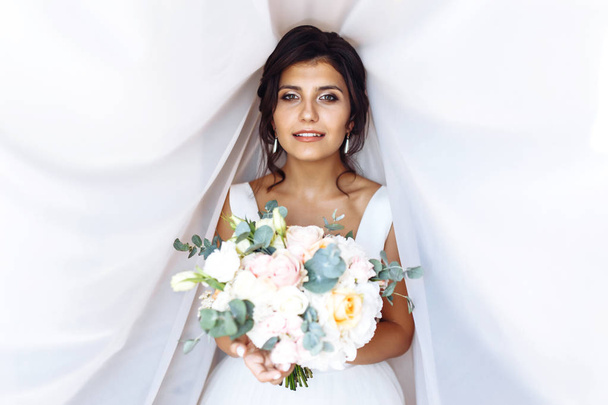 Young bride in white dress with a bouquet of flowers posing under curtain. Beautiful woman with professional make up and hair style. Morning of the bride. Wedding day. Fashion bride. - Foto, immagini