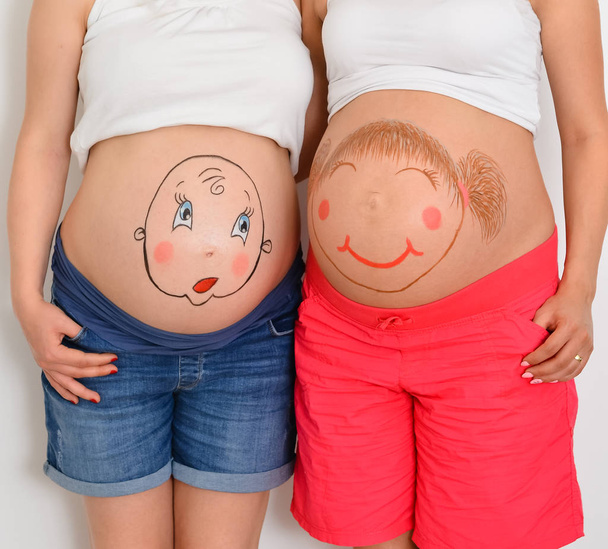 Premier accouchement. Gros plan ventre de deux jeunes femmes enceintes caucasiennes avec visage d'enfants souriants ventre peint debout contre un mur blanc.
  - Photo, image