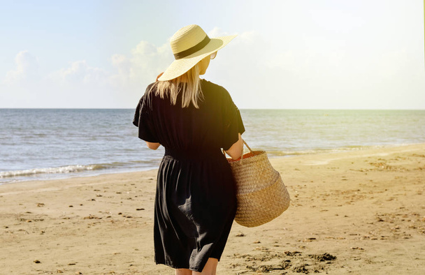 Rear view of young  caucasian woman with dress  ,  straw hat and basket waliking  on beach   and relaxing . Summer holidays . - Photo, Image