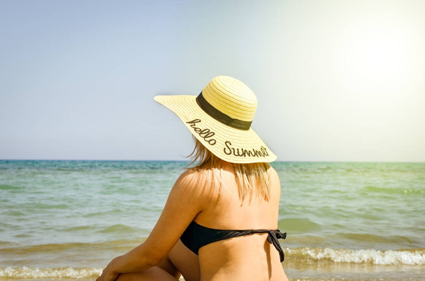 Блондинка одягнена в чорні купальники і великий солом'яний капелюх на голові, який засмагає і розслабляється на березі моря .. Яскравий теплий фільтр
 . - Фото, зображення