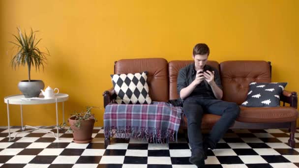 Mann spielt Spiel am Telefon auf der Couch sitzend. Archivmaterial. Junge Mann spielt begeistert Telefon sitzen in stilvollem Interieur. Junger Mann verbringt Freizeit mit Spielen am Telefon - Filmmaterial, Video