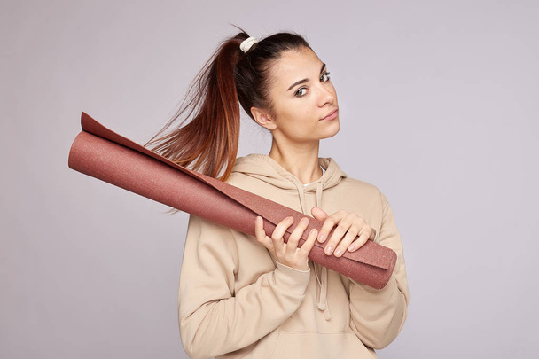 Sport concept. Dark haired young fitness woman holds rolled rubber mat, looks at camera, ready for doing exercises indoor, dressed in sportswear. Athletic attractive female prepares for yoga training. - 写真・画像