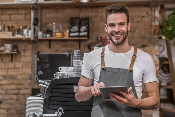 Улыбающийся молодой предприниматель в фартуке рядом со стойкой кафе и с цифровым планшетом
 - Фото, изображение