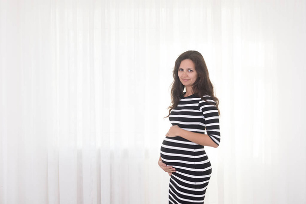Pregnant woman in striped dress holds hands on belly on a white background. Pregnancy, maternity, preparation and expectation concept. Beautiful tender mood photo of pregnancy. - Photo, Image
