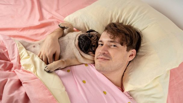 Handsome man lying with pug at bed. Young man in a bed under a rug with his dog in an embrace. Handsome young hipster with his pug resting on bed at home - Photo, image