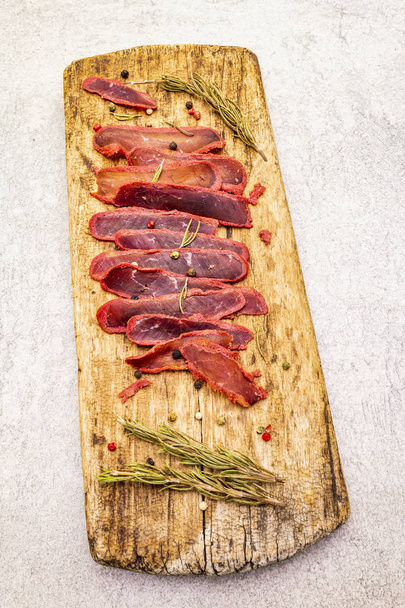 Basturma, dried tenderloin of beef meat, jerky, thinly sliced. Dry rosemary, pepper mix on vintage wooden board. Delicious food on a stone background. - Photo, Image