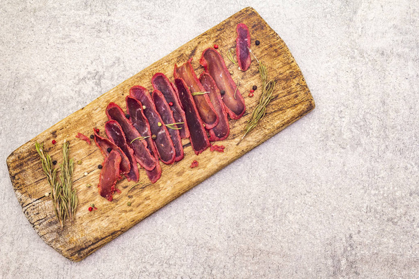 Basturma, dried tenderloin of beef meat, jerky, thinly sliced. Dry rosemary, pepper mix on vintage wooden board. Delicious food on a stone background, top view, copy space. - Photo, Image