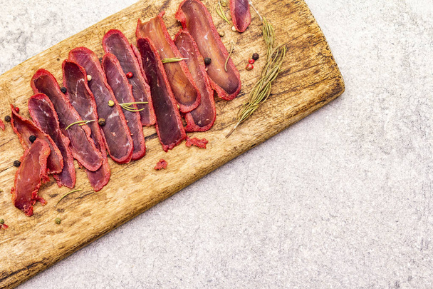 Basturma, dried tenderloin of beef meat, jerky, thinly sliced. Dry rosemary, pepper mix on vintage wooden board. Delicious food on a stone background, top view, copy space, close up. - Foto, Imagem