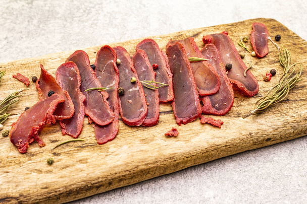 Basturma, dried tenderloin of beef meat, jerky, thinly sliced. Dry rosemary, pepper mix on vintage wooden board. Delicious food on a stone background, close up. - Photo, Image
