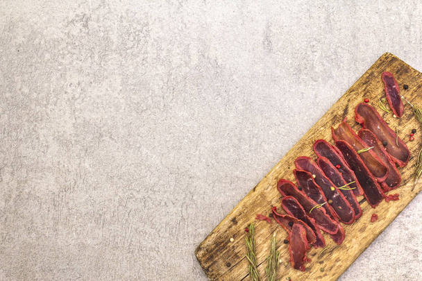 Basturma, dried tenderloin of beef meat, jerky, thinly sliced. Dry rosemary, pepper mix on vintage wooden board. Delicious food on a stone background, top view, copy space. - Photo, Image