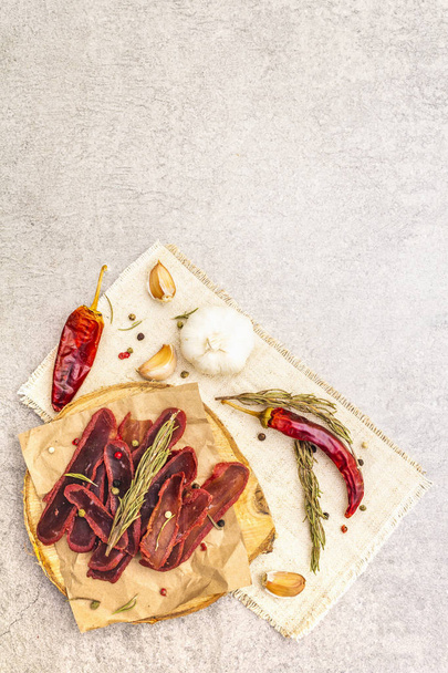 Basturma, dried tenderloin of beef meat, jerky, thinly sliced. Dry rosemary, pepper mix, chili, garlic on vintage linen cloth. Delicious food on a wooden board, stone background, copy space, top view. - Photo, Image