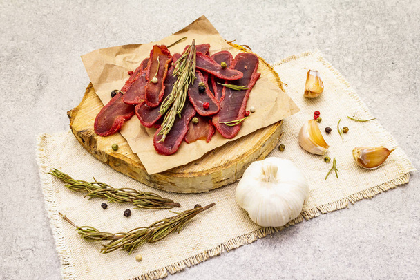 Basturma, dried tenderloin of beef meat, jerky, thinly sliced. Dry rosemary, pepper mix, chili, garlic on vintage linen cloth. Delicious food on a wooden board, stone background, close up. - Photo, Image