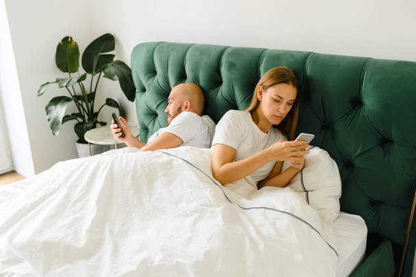 young couple lie in bed with their phones and turned away from each other - Foto, Imagem