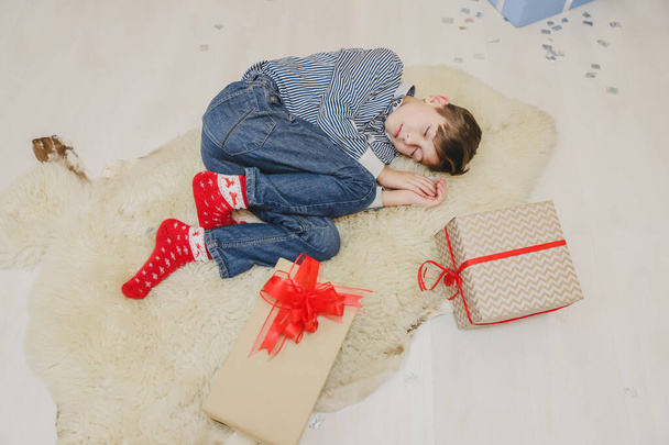 Lovely image of little boy sleeping on white carpet, among Christmas presenst, curling up. - Foto, imagen