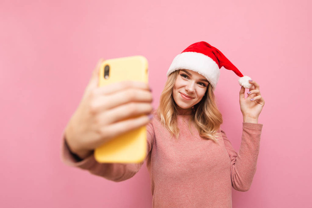Ragazza attraente in cappello di Babbo Natale fa selfie su sfondo rosa, guarda nella fotocamera di smarphone e pose. Ragazza positiva in cappello di Natale fa foto di vacanza sulla fotocamera dello smartphone. Isolato
. - Foto, immagini