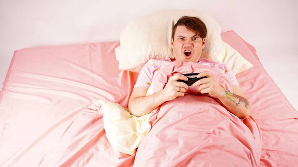 Чоловік з джойстиком грає в відеогру, лежачи на ліжку. Молодий чоловік з джойстиком, концепція ігрової залежності, домашнє дозвілля
 - Фото, зображення