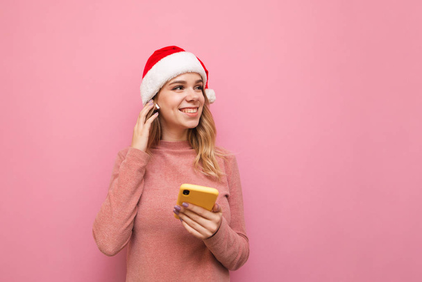 Attractive happy girl in Christmas and hat stands with smartphone in hand, listens to music in wireless headphones, smiles and looks away at copyspace. Isolated on pink background. Christmas concept. - Photo, Image