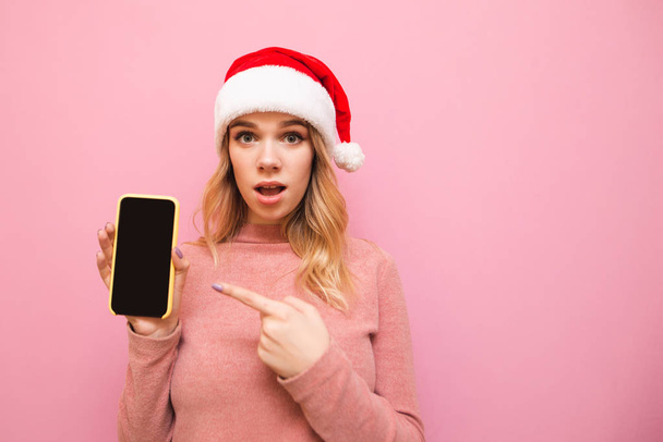 Megdöbbent lány egy karácsonyi kalap áll egy rózsaszín háttér, tartja az okostelefon a kezében, mutatja az ujját a képernyőn. Megdöbbent hölgy ujjal mutogat egy okostelefonra, és a kamerába néz. - Fotó, kép