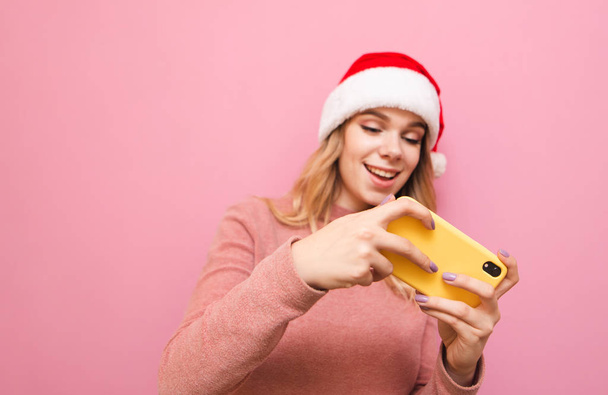 ゲーマーの女の子の手の中にスマートフォンに焦点を当てます。クリスマスの帽子の正のティーンブロンドの女の子は、ピンクの背景に隔離された画面と笑顔で、スマートフォンでモバイルゲームをプレイします - 写真・画像