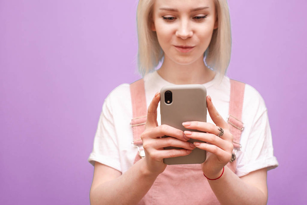 Smartphone en manos de una adolescente. Foto de cerca de una chica rubia en ropa linda utiliza un teléfono inteligente sobre un fondo púrpura, el enfoque en el teléfono en las manos de la chica. Espacio de copia
 - Foto, imagen