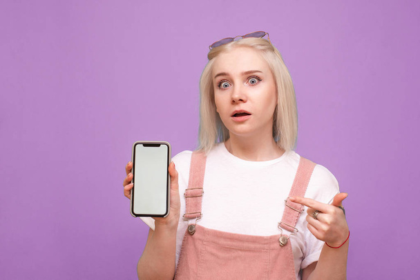 Surprised girl wears a light casual clothing, holds a smartphone with a white screen, shows a finger on the screen and looks into the camera.Teen girl shows a finger on a phone in hand,surprised face - Foto, Imagen