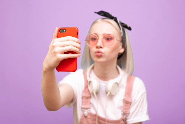 Κοντινό πλάνο πορτρέτο ενός αστείου κοριτσιού με έντονα ροζ γυαλιά βγάζει μια selfie σε ένα smartphone με ένα αστείο πρόσωπο σε μωβ φόντο. Εστίαση στο smartphone. - Φωτογραφία, εικόνα