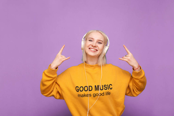Η χαρούμενη ξανθιά με πορτοκαλί μπλούζα και ακουστικά είναι σε μωβ φόντο, ακούει μουσική και δείχνει ένα σημάδι heavy metal, κοιτάζει την κάμερα και χαμογελάει. Μεμονωμένα - Φωτογραφία, εικόνα