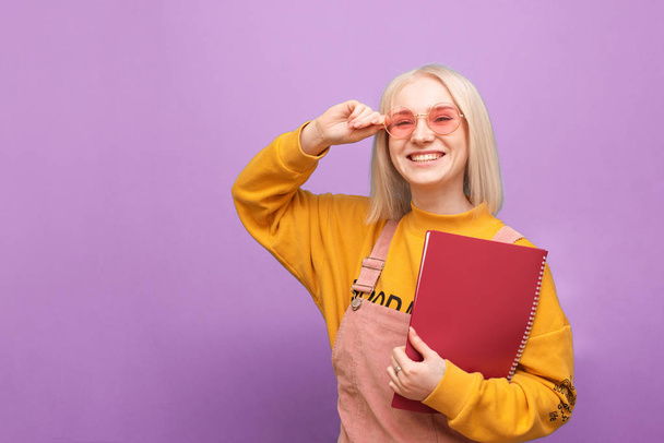 Retrato de una estudiante feliz con gafas de color rosa y un bloc de notas en la mano de pie sobre un fondo morado, mira a la cámara y sonríe. Chica con estilo con libros en sus manos sonriendo, aislado
. - Foto, imagen