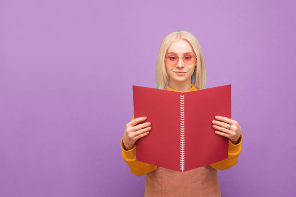 Portrait of a funny blonde in pink glasses on a purple background, holds a red notebook in his hands and reads. Cheerful girl student is learning. Isolated. - Foto, Imagem