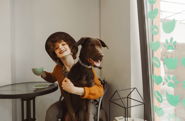 A happy girl in a hat sits in a cafe with a cup of coffee and a dog on her lap, looks into the camera and smiles. The owner with a pet in her hands is having breakfast at the cafe. - Foto, Bild